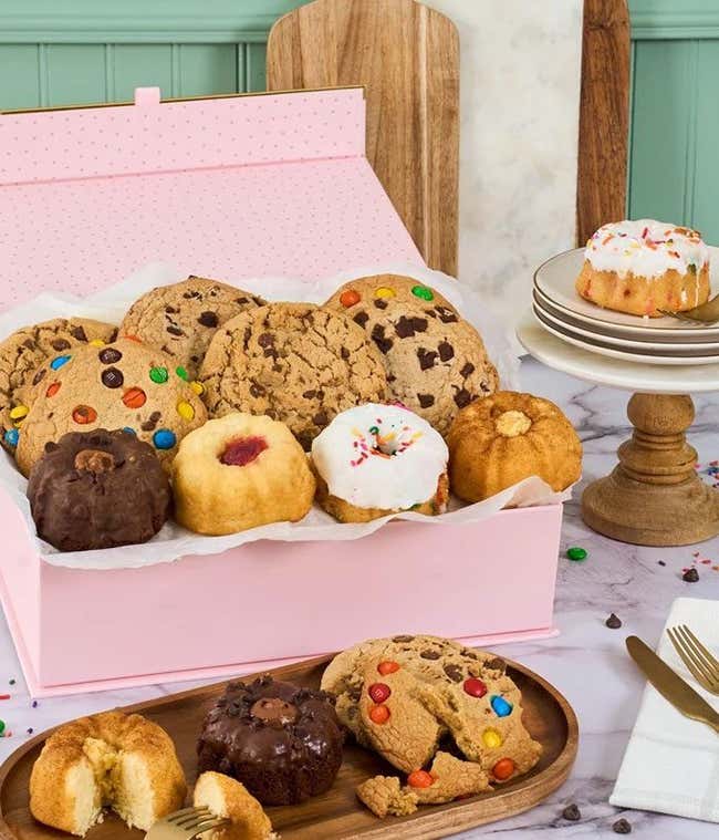 Two rainbow striped gift boxes with a blue bow. In front, a plate with a mini bundt cake with a candle, another tray with cookies, brownies, and blondies. Behind those, a cake tray with whoopie pies and mini bundt cakes. 