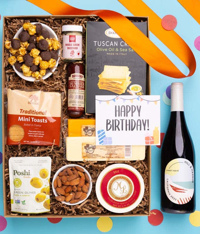 A charcuterie gift set with Tuscan crisps, focaccia crackers, 3 cheeses, summer sausage, olives, mustard, smoked almonds, chocolate-covered popcorn, and a birthday card.