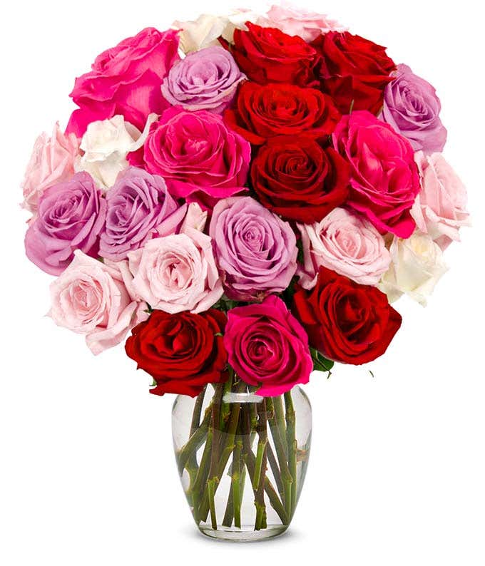 A bouquet of red, pink, lavender, and white roses with green leaves arranged in a clear glass vase.