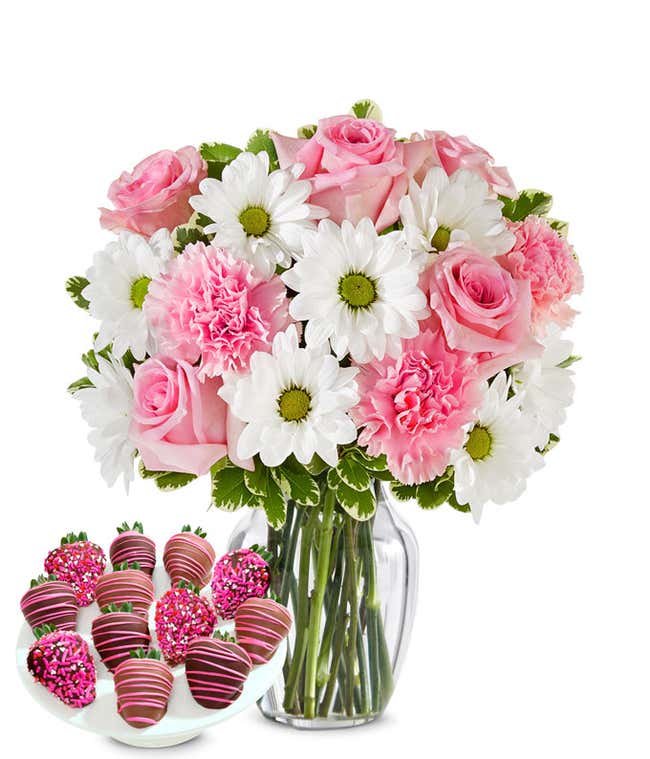 Charming bouquet of pink roses, pink carnations, and white daisies in a clear glass vase, paired with chocolate-covered strawberries decorated with pink drizzle and pink sprinkles.