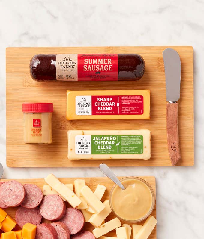 Two wooden boards with sliced summer sausage, cubes of sharp and jalapeño cheddar cheese, sweet hot mustard, and cheese spreaders, with the full sausage, cheese blocks, and mustard jar displayed above.