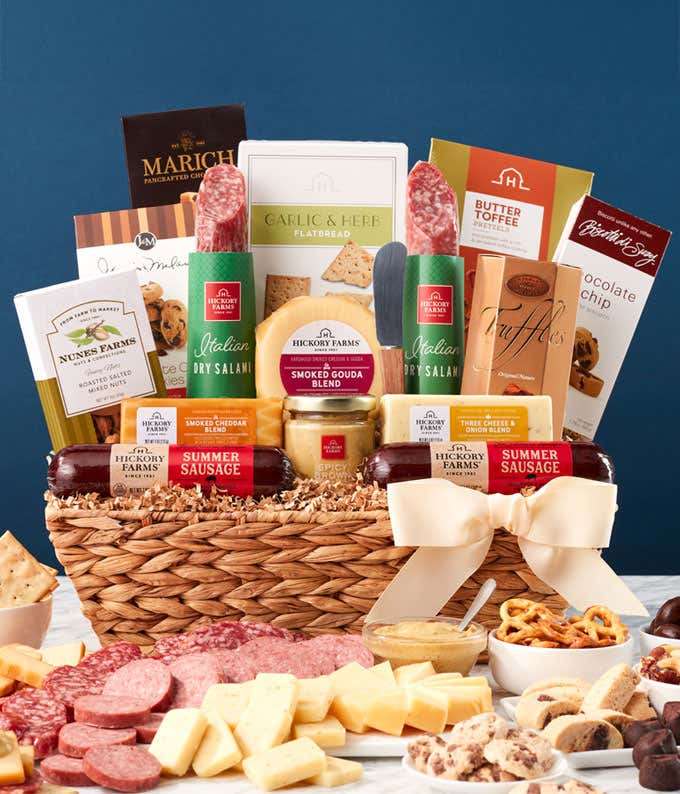 Woven gift basket filled with summer sausages, salami, assorted cheeses, crackers, mustards, cookies, chocolate truffles, and snacks. A serving board in front displays sliced sausage, cheese wedges, mustard, and pretzels, with a white bow on the basket.