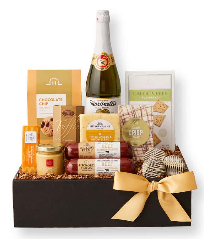 Gift basket with sparkling cider, chocolate chip cookies, crackers, cheese spreads, sausage, garlic &amp; herb flatbread, and truffles in a black box with a gold bow, surrounded by a serving of sausage, cheese, and crackers on a marble surface.
