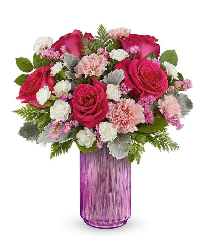 Vibrant floral arrangement featuring 50cm hot pink roses, pink carnations, miniature white carnations, raspberry sinuata statice, dusty miller, leatherleaf fern, and lemon leaf in a textured pink glass vase.