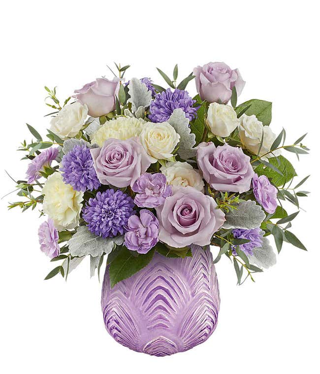 Elegant floral arrangement featuring lavender roses, light yellow carnations, Cream Spray roses, purple asters, dusty miller, and lush greenery in a textured lavender vase with a chevron-inspired design