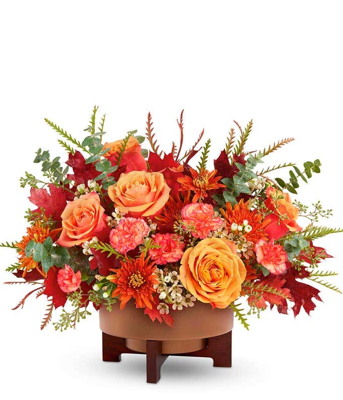 Fall flower arrangement with peach roses, orange carnations, rust-colored daisy chrysanthemums, white waxflower, and assorted autumn leaves in a brown ceramic container on a wooden stand.