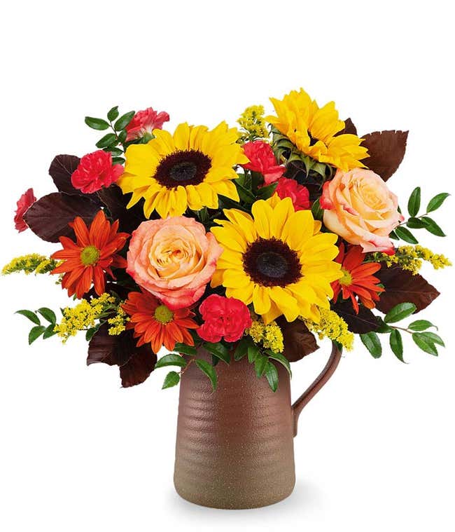 Fall floral arrangement with yellow sunflowers, orange bi-color roses, red spray roses, orange daisy spray chrysanthemums, and yellow solidago in a brown ceramic pitcher.