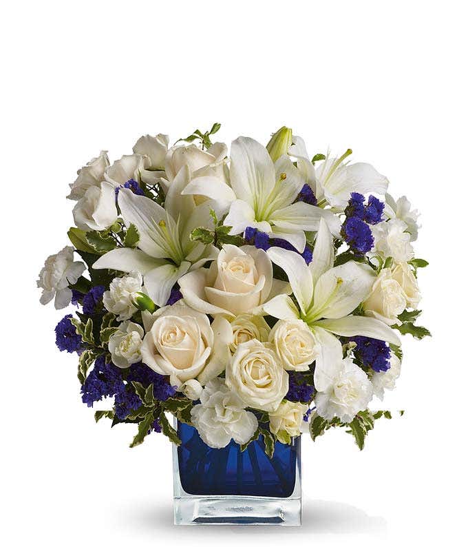 Floral arrangement featuring crème roses, crème spray roses, white Asiatic lilies, white miniature carnations, and purple statice in a blue cube vase.