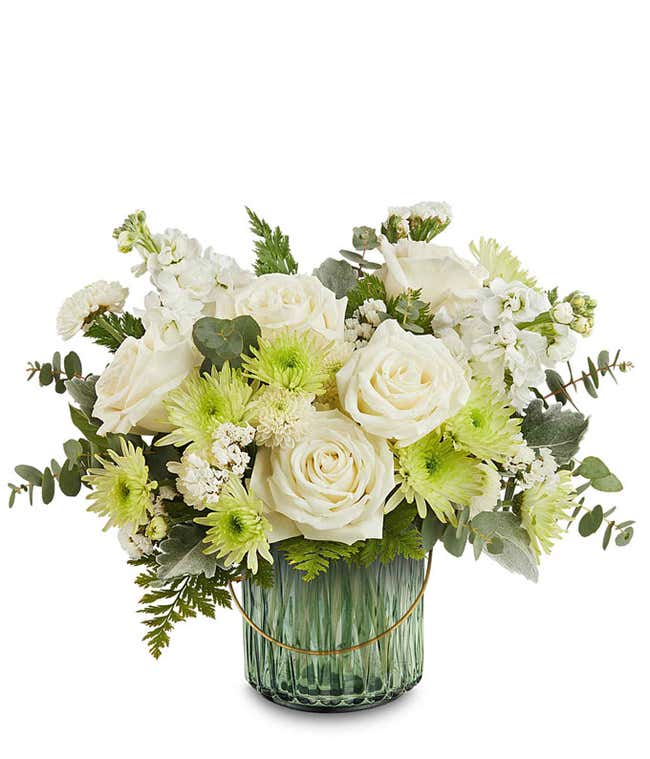 White roses, white stock, green button spray chrysanthemums, white cushion spray chrysanthemums, and assorted greenery arranged in a ribbed green glass vase