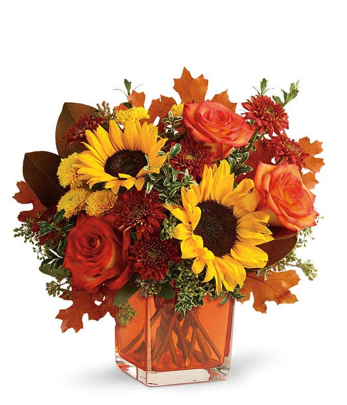 A vibrant floral arrangement featuring orange roses, yellow sunflowers, gold cushion poms, and rust cushion mums, accented with seeded eucalyptus, oregonia, and magnolia leaves, all beautifully presented in a keepsake orange cube vase.