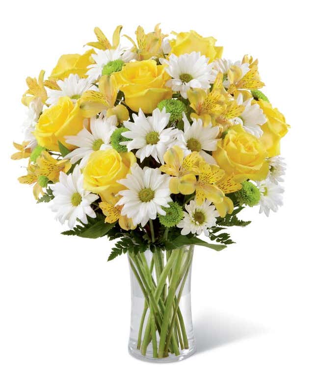 Bright bouquet featuring yellow roses, yellow alstroemeria, white daisies, green poms, and assorted greenery arranged in a tall clear glass vase. 