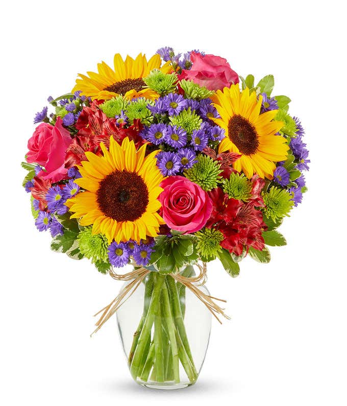 Bright mixed flower bouquet featuring yellow sunflowers, hot pink roses, red alstroemeria, green button poms, and purple asters in a clear glass vase tied with raffia ribbon.