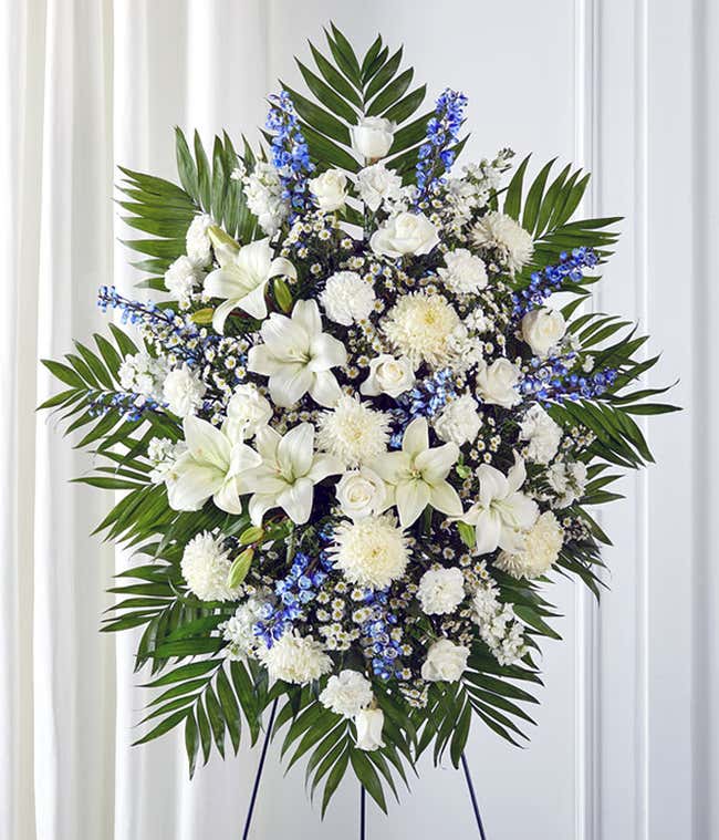 Standing spray arrangement featuring white lilies, roses, carnations, and poms, accented with blue delphinium, white monte casino, and palm fronds on a display easel. 