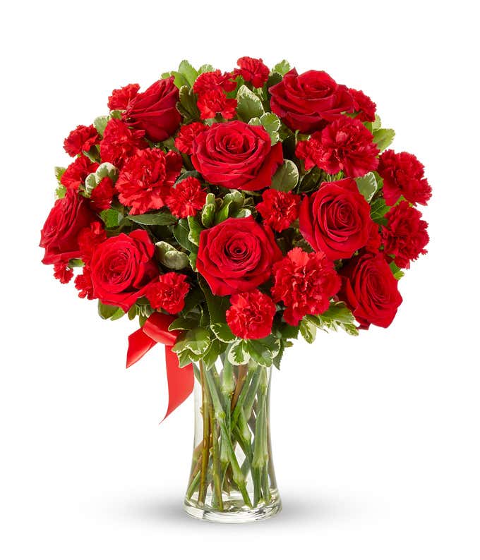 A bouquet of red roses and red carnations with variegated greenery and a red ribbon in a clear glass vase.
