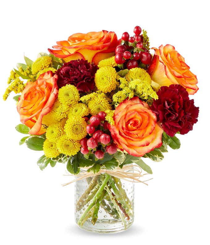 Small clear glass jar filled with orange roses, burgundy carnations, bright yellow button poms, red berries, and greenery, tied with a raffia bow.