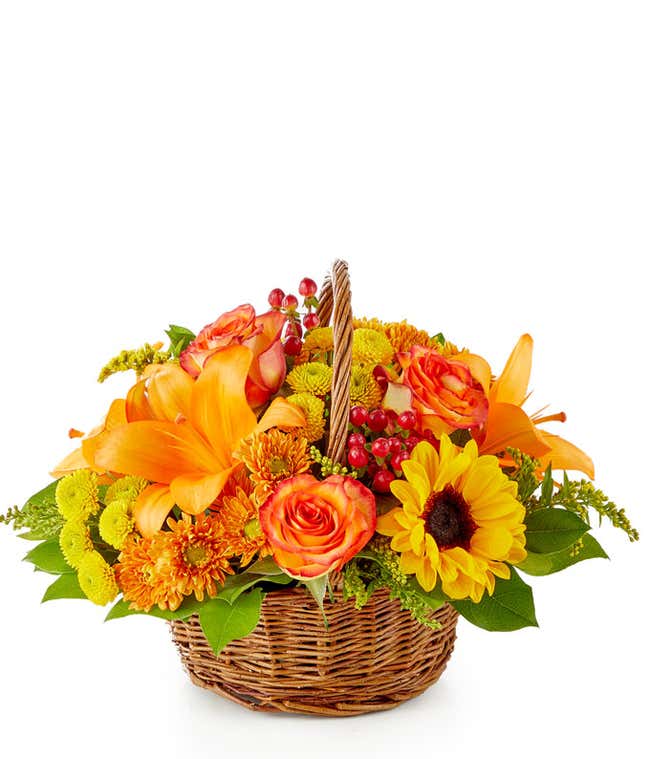 Woven basket filled with a vibrant autumn-inspired floral arrangement featuring orange lilies, bi-color orange roses, yellow sunflowers, bronze chrysanthemums, red hypericum berries, green button poms, yellow solidago, and assorted greenery.