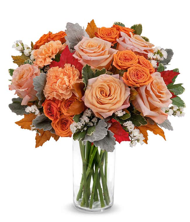 Clear glass vase filled with a lush arrangement of peach and orange roses, orange carnations, white filler flowers, dusty miller, red and orange autumn leaves, and assorted greenery.