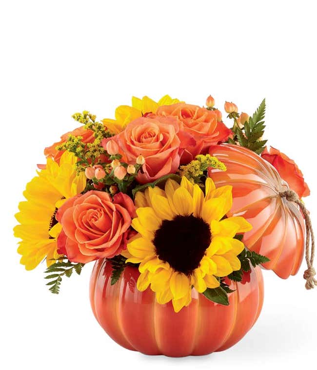 Fall flower arrangement with orange roses, sunflowers, yellow solidago, peach hypericum berries, and greenery in a ceramic pumpkin container with a lid and rope accent.