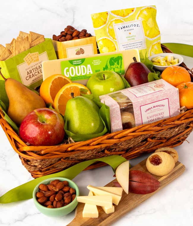 A basket filled with fresh fruits, crackers, cheese crisps, almonds, honey mustard, and assorted caramels, adorned with a green ribbon. 