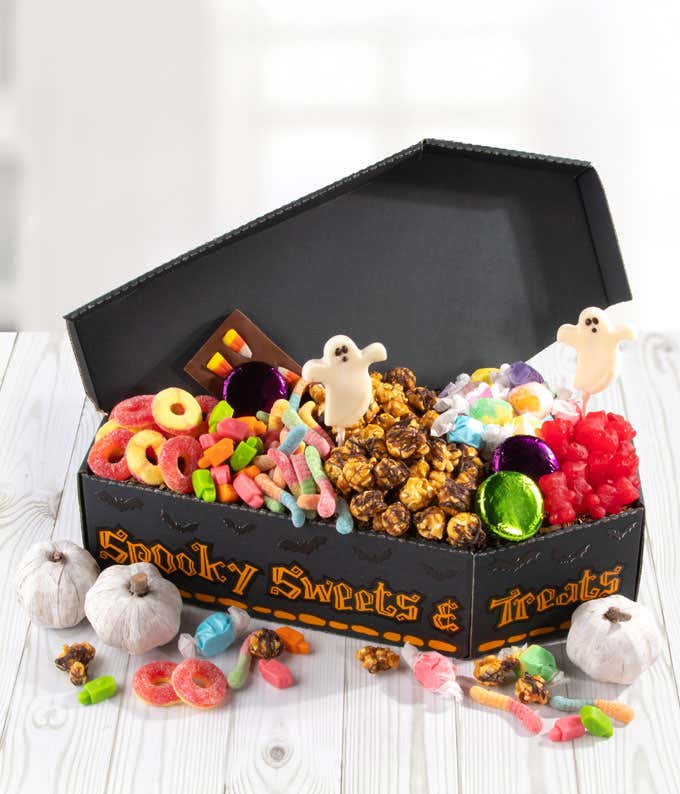Halloween coffin-shaped gift box filled with chocolate-covered popcorn, ghost lollipops, gummy candies, foil-wrapped chocolates, salt water taffy, and assorted sweets, with decorative mini pumpkins and scattered candies around the box.