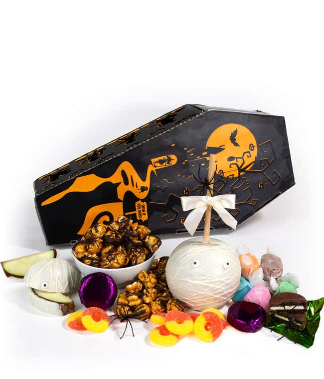 Halloween coffin-shaped gift box with a mummy design, filled with chocolate-covered popcorn, foil-wrapped chocolates, salt water taffy, gummy candies, and a white chocolate-covered apple decorated like a mummy, surrounded by plastic spiders.