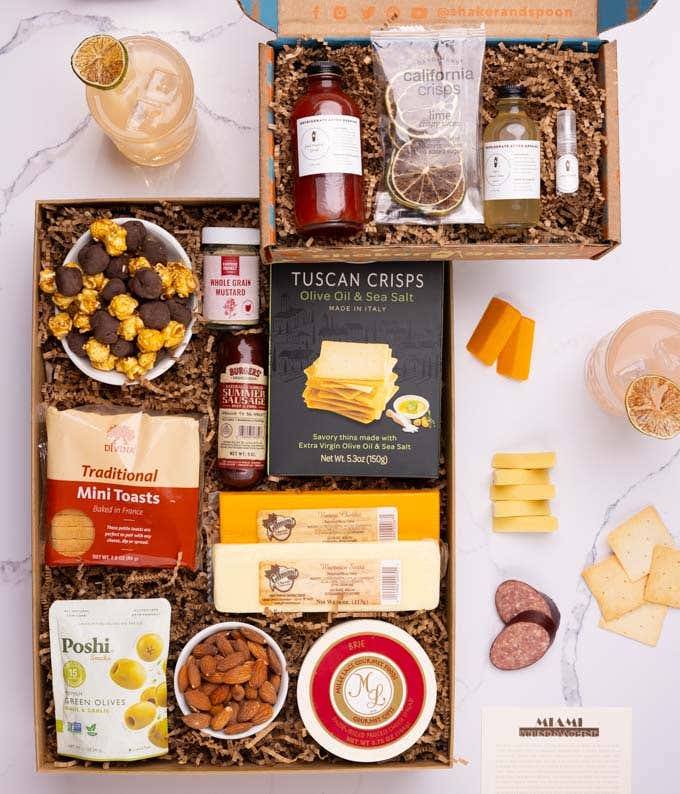 The food box contains three cheeses, summer sausage, crackers, almonds, olives, popcorn and mustard.  The cocktail box contains tropical pink syrup, lemon-lime juice, rose water, dehydrated lime slices, and recipe card.