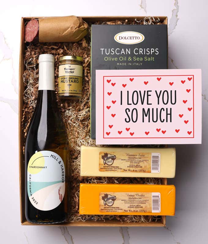 A beautifully arranged gift box featuring a bottle of Hill & Harbor Pinot Noir, two hard cheeses, a jar of stone-ground mustard, gourmet crackers, savory summer sausage, and a pink card with hearts that reads 'I Love You So Much'