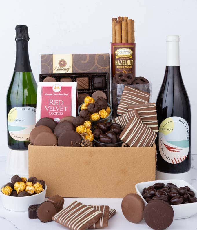 A gift box overflowing with treats including an assortment of chocolate-covered items like oreoes, almonds graham crackers and pretzels along with red velvet cookies, hazelnut wafers, an 8 piece box of chocolates and 2 bottles of wine.