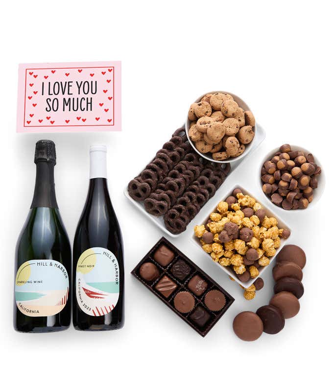 A gift box overflowing with treats including an assortment of chocolate-covered items like oreoes, almonds graham crackers and pretzels along with red velvet cookies, hazelnut wafers, an 8 piece box of chocolates, 2 bottles of wine and an I Love You card.