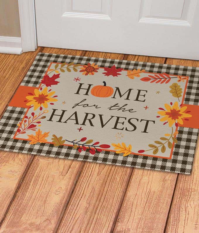 Home for the Harvest doormat with autumn-themed design, featuring fall leaves and a pumpkin, bordered by a black and white buffalo plaid pattern.